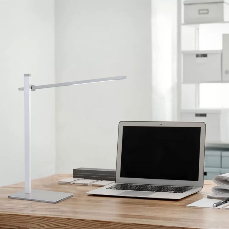 Ein moderner Arbeitsplatz mit einer eleganten, silbernen Tischleuchte - Leve und einem offenen Laptop auf einem Holzschreibtisch. Im Hintergrund sind weiße Regale mit ordentlich angeordneten Kisten zu sehen.