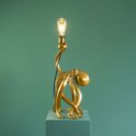 Die Tischleuchte - Oktopus ist eine dekorative Lampe in Form eines goldenen Oktopus mit einer freiliegenden Glühbirne im Vintage-Stil, die elegant auf einem blaugrünen Sockel vor einem passenden blaugrünen Hintergrund thront.