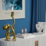 Eine goldene Tischleuchte - Oktopus mit freiliegender Glühbirne steht auf einem weißen Schrank. Daneben sorgen gläserne Kerzenständer und Bücher für Charme. Korallenfarbenes Dekor ergänzt ein gerahmtes „Bondi Beach“-Poster an der blauen Wand und schafft eine einladende Küstenatmosphäre.