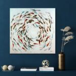 Das Wandbild – Fische ziert eine blaue Wand über einem Regal mit Dekokugeln und gestapelten Büchern, ergänzt durch eine hohe, goldene Vase mit Trockenpflanzen auf der rechten Seite.