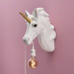 Die Wandleuchte „Unicorn“ besteht aus einem weißen Einhornkopf mit goldenem Horn und einer Glühbirne, die an einem weißen Kabel aus seinem Mund hängt. Sie ist an einer rosa Wand angebracht und verbreitet ein warmes Licht.