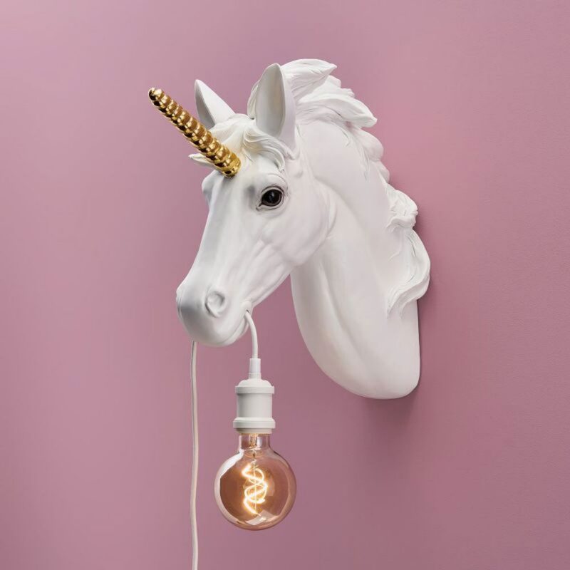 Die Wandleuchte „Unicorn“ besteht aus einem weißen Einhornkopf mit goldenem Horn und einer Glühbirne, die an einem weißen Kabel aus seinem Mund hängt. Sie ist an einer rosa Wand angebracht und verbreitet ein warmes Licht.