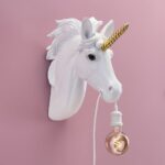 Die Wandleuchte - Einhorn mit einem weißen Einhornkopf mit goldenem Horn und wallender Mähne ist an einer rosa Wand montiert. Sie hält elegant eine hängende Glühbirne mit einem weißen Kabel im Mund.