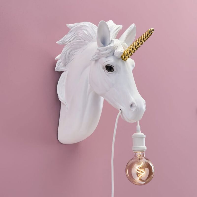Die Wandleuchte - Einhorn mit einem weißen Einhornkopf mit goldenem Horn und wallender Mähne ist an einer rosa Wand montiert. Sie hält elegant eine hängende Glühbirne mit einem weißen Kabel im Mund.