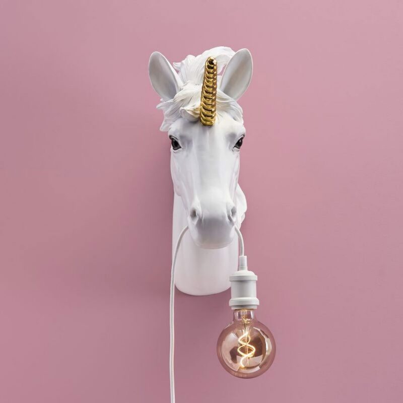 Die weiße Wandleuchte - Wanddekoration Einhorn mit goldenem Horn ist an einer rosa Wand montiert. Sie scheint eine leuchtende Glühbirne am Mund zu halten, die elegant an einem weißen Kabel hängt.