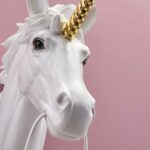 Diese skurrile Wandleuchte „Unicorn“ zeigt eine Nahaufnahme einer weißen Einhornbüste mit einem glänzenden goldenen Horn vor einem rosa Hintergrund. Ein Seil in seinem Mund verleiht dem Kunstwerk eine bezaubernde Note.