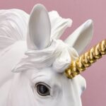 Eine Nahaufnahme der Wandleuchte - Einhorn-Skulptur zeigt einen weißen Körper und eine detaillierte Mähne vor einem rosa Hintergrund. Sein spiralförmiges goldfarbenes Horn verleiht Charme und große, realistische Augen vermitteln Laune und Fantasie.