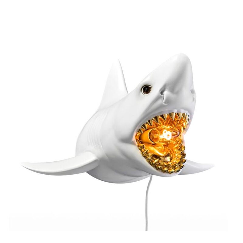 Die Wandleuchte - Weißer Hai ist eine weiße Wandleuchte in Form eines Haikopfes mit offenem Maul, der eine goldgelbe Glühbirne freigibt, die von Zähnen umgeben ist und so einen beleuchteten Kiefereffekt erzeugt. Ein Netzkabel verläuft von der Rückseite.