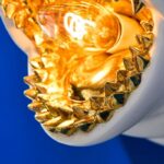 Eine Nahaufnahme der Wandleuchte – Weißer Hai zeigt eine leuchtende Glühbirne, die von dekorativen goldenen Spitzen umgeben ist, vor einem hellblauen Hintergrund.