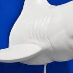 Nahaufnahme der glatten, stilisierten Rückseite der „Wandleuchte – Weißer Hai“, die einem Hai oder Fisch mit Flossendetails ähnelt und als kunstvolle Lampe vor einer leuchtend blauen Wand dient.