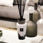 Der Baobab Diffusor - Platinum mit langen schwarzen Stäbchen steht auf einem Glastisch. Verschwommene graue Kissen und runde dekorative Glasobjekte im Hintergrund tragen zum gemütlichen, modernen Ambiente bei.