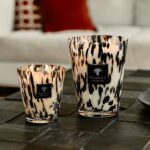 Zwei Kerzen aus der Baobab Diffuser- Platinum Collection mit schwarz-weiß gesprenkeltem Design auf Glashaltern leuchten auf einem dunklen Tisch. Im Hintergrund liegt ein rotes Kissen auf einem leicht verschwommenen Sofa. Etiketten auf den Haltern weisen auf ihre Herkunft hin.