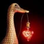 Eine goldene Entenskulptur hält die leuchtende Glühbirne Leuchtmittel - Red Heart anmutig in ihrem Schnabel. Die rote Glühbirne hängt an einem schwarzen Kabel mit einer Messinghalterung und erzeugt ein beeindruckendes Bild vor einem dunklen Hintergrund, der ihr lebendiges Leuchten verstärkt.