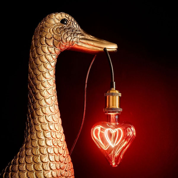 Eine goldene Entenskulptur hält die leuchtende Glühbirne Leuchtmittel - Red Heart anmutig in ihrem Schnabel. Die rote Glühbirne hängt an einem schwarzen Kabel mit einer Messinghalterung und erzeugt ein beeindruckendes Bild vor einem dunklen Hintergrund, der ihr lebendiges Leuchten verstärkt.