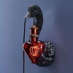 Eine schwarze, schwanenförmige Wandskulptur hält auf elegante Weise ein „Leuchtmittel – Red Heart“, eine herzförmige rote Glasbirne mit sichtbarem Glühfaden, vor der blauen Wand und verbindet sich nahtlos mit einer metallischen Fassung am Schnabel des Schwans.