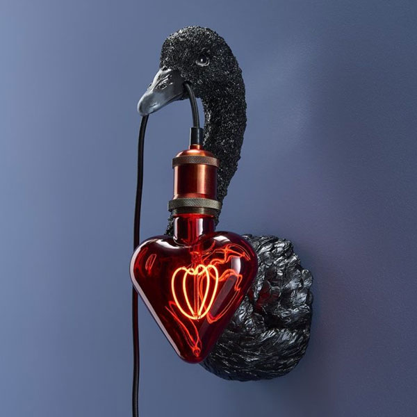 Eine schwarze, schwanenförmige Wandskulptur hält auf elegante Weise ein „Leuchtmittel – Red Heart“, eine herzförmige rote Glasbirne mit sichtbarem Glühfaden, vor der blauen Wand und verbindet sich nahtlos mit einer metallischen Fassung am Schnabel des Schwans.