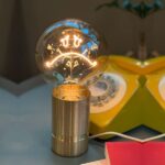 Eine Statement-Glühbirne - Doppelherz Warmweiß mit einem leuchtenden traurigen Gesicht sitzt auf einem metallischen Sockel. Dahinter ruht auf einer blaugrauen Oberfläche ein gelbes Vintage-Telefon, im Vordergrund ist ein kleiner Teil eines offenen rot-weißen Buches zu sehen.