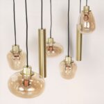Die Pendelleuchte - Lucy 2.0 - 9er verfügt über bernsteinfarbene Glasschirme mit zylindrischen Messingbefestigungen, die in unterschiedlichen Höhen hängen und freiliegende Glühlampen zur Schau stellen, die für ein warmes, stilvolles Ambiente sorgen.