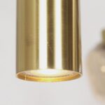 Nahaufnahme einer modernen zylindrischen Pendelleuchte aus Messing - Lucy 2.0 - 9er mit glatter Oberfläche. Das Licht ist eingeschaltet und wirft aus seiner offenen Unterseite ein sanftes Licht vor einem unscharfen Hintergrund, was seine Eleganz betont.