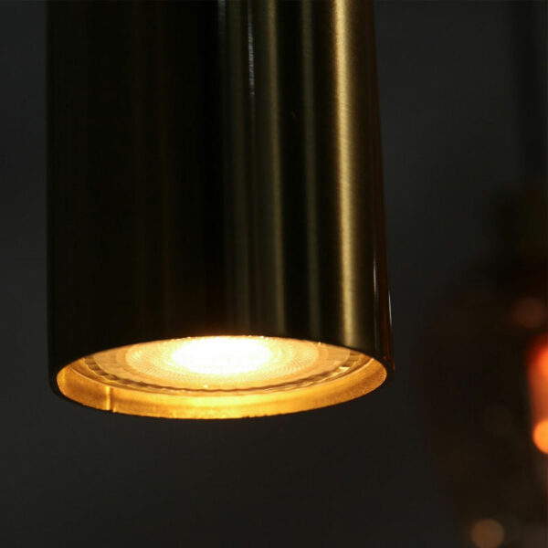 Die Pendelleuchte - Lucy 2.0 - 9er wirft einen warmen Schein auf einen dunklen Hintergrund, wobei ihre glatte, glänzende Metalloberfläche das Licht sanft reflektiert.