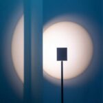 Ein schwarzes rechteckiges Objekt wirft Schatten auf ein großes weißes, rundes Licht, das auf eine blaue Wand projiziert wird und an das minimalistische Design der Stehleuchte - Liva erinnert. Das Zusammenspiel von Licht und Schatten erzeugt ein abstraktes Bild.