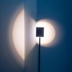 Die Stehleuchte - Liva wirft Schatten auf eine Eckwand und bildet überlappende Kreisformen. Ein quadratisches Objekt auf einem Ständer blockiert das Licht teilweise und erzeugt so ein künstlerisches Muster vor dem blauen Hintergrund.