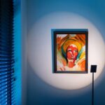 Eine abstrakte Gesichtsbemalung in leuchtendem Rot, Orange und Blau ziert eine Wand. Der schwach beleuchtete Raum hebt das Kunstwerk mit einem Strahler der Stehleuchte - Liva hervor, während vertikale Jalousien auf der linken Seite einen Hauch von blauem Licht verbreiten.