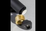 Nahaufnahme von zwei Pendelleuchten - Puc 10er auf einer grauen Oberfläche. Eine ist offen mit einem weißen Element und zeigt ein mattschwarzes Äußeres. Daneben befinden sich zwei Kappen, eine in Metallic-Gold und die andere in Metallic-Grau.