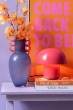 Eine lila Vase mit orangen Blumen steht neben der Tischleuchte – Toad pink auf einem Regal. Unter der Lampe liegen zwei gestapelte Bücher, „Plant Tribe“ und „Nordic Moods“. Als Hintergrund dient ein leuchtend oranges Poster mit pinkem Text.