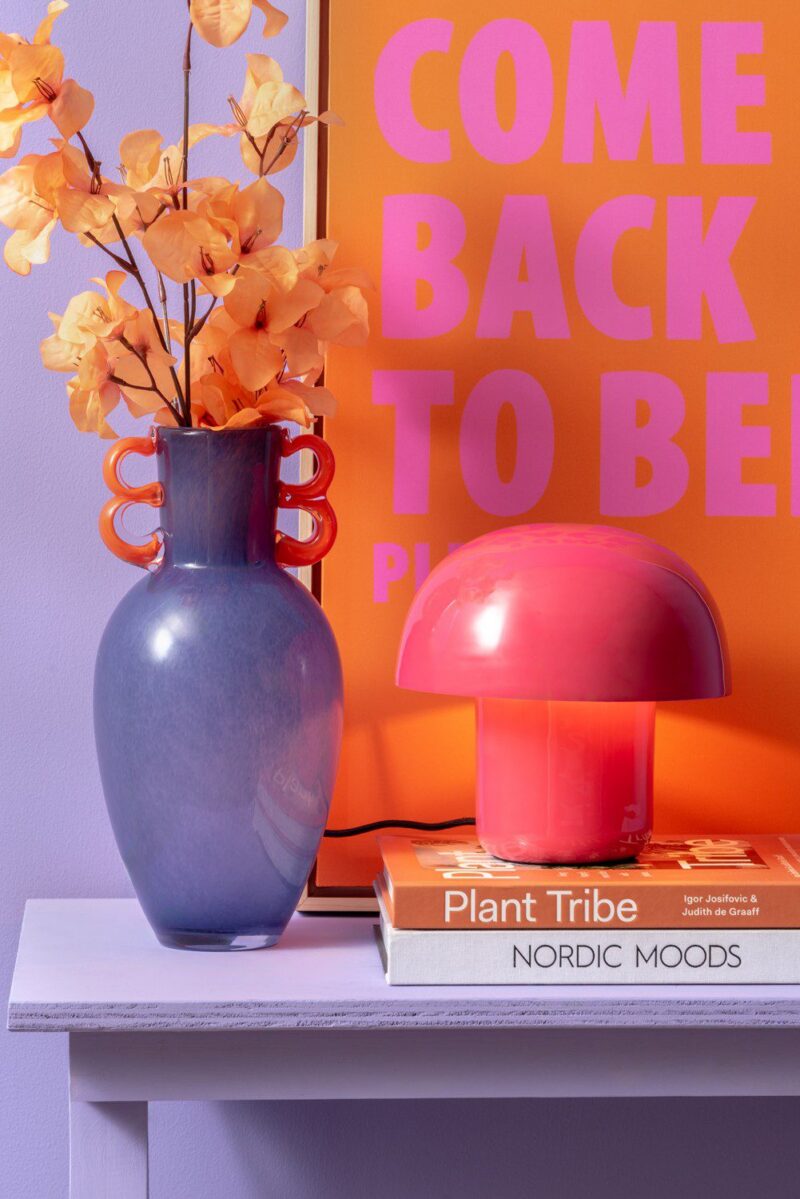 Eine lila Vase mit orangen Blumen steht neben der Tischleuchte – Toad pink auf einem Regal. Unter der Lampe liegen zwei gestapelte Bücher, „Plant Tribe“ und „Nordic Moods“. Als Hintergrund dient ein leuchtend oranges Poster mit pinkem Text.