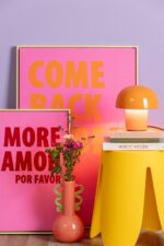 In einem lebhaften Raum hängen zwei gerahmte rosa Poster mit den Aufschriften „KOMM ZURÜCK“ und „MORE AMOR POR FAVOR“. Auf einem gelben Tisch stehen Bücher und eine orange Tischleuchte – Toad-Lampe. Vor den Postern stehen rosa Blumen in einer pfirsichfarbenen Vase, alles vor einer violetten Wand.
