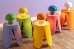 Eine Reihe von fünf farbenfrohen „Tischleuchten - Toad“-Lampen steht auf passenden Hockern in Gelb, Lila, Grün, Orange und Pink. Sie stehen auf einem Holzboden vor einer lavendelfarbenen Wand.