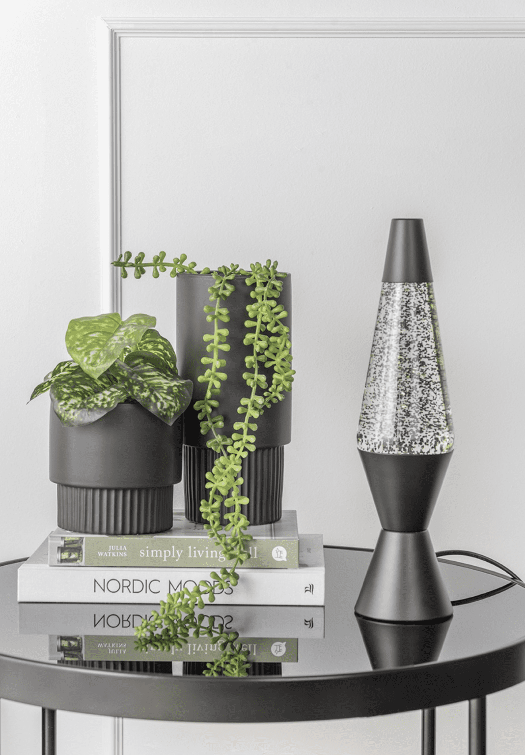 Auf einem kleinen schwarzen Tisch stehen zwei schwarze Blumentöpfe mit grünen Blattpflanzen, gestapelte Bücher mit den Titeln „Simply Living“ und „Nordic Moods“ und eine schwarze Tischlampe – Lava Glitter. Der Hintergrund ist eine weiße Wand.