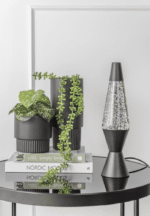 Auf einem kleinen schwarzen Tisch stehen zwei schwarze Blumentöpfe mit grünen Blattpflanzen, gestapelte Bücher mit den Titeln „Simply Living“ und „Nordic Moods“ und eine schwarze Tischlampe – Lava Glitter. Der Hintergrund ist eine weiße Wand.