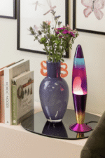 Eine Tischleuchte Lava Multicolor und eine violette Vase mit Blumen stehen auf einer verspiegelten Oberfläche. Daneben sind Bücher wie „Nordic Moods“ und „Einfach gut leben“ zu sehen. Im Hintergrund ist Wandkunst mit Schmetterlingsmotiven zu sehen.