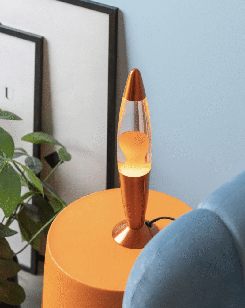 Eine leuchtende Tischleuchte Lava Multicolor steht auf einem orangefarbenen Tisch neben einem blauen Sofa. Im Hintergrund stehen eine Zimmerpflanze und gerahmte Bilder vor einer hellblauen Wand.