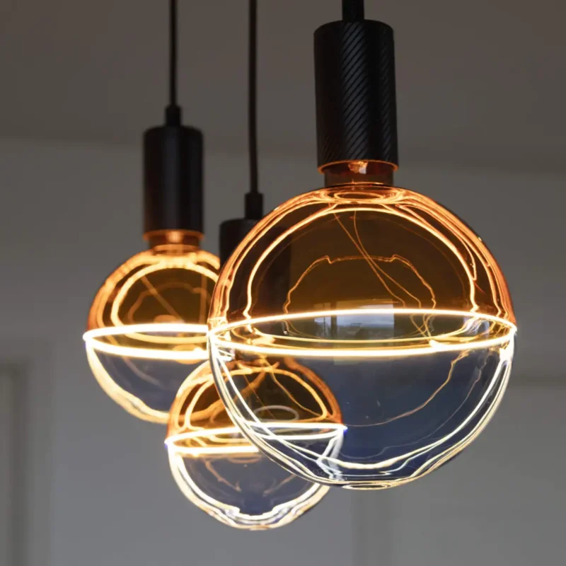 Drei kugelförmige LED-Pendelleuchten Leuchtmittel-Illusion von Licht Factory hängen von der Decke. Mit ihrem Filamentdesign und den schwarzen Sockeln verbreiten diese Leuchten ein warmes, goldenes Licht und schaffen vor einem neutralen Hintergrund eine stilvolle und gemütliche Atmosphäre.