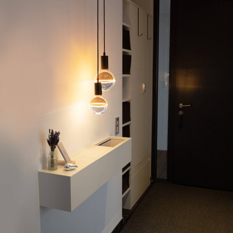 Ein moderner Flur mit einer eleganten schwarzen Tür und zwei Illusion LED-Leuchtmitteln von Licht Factory, die ein warmes Licht verbreiten. Eine minimalistische weiße Konsole zeigt eine Vase mit Lavendel und eine kleine Schale, während die Wand mit offenen Regalen ausgestattet ist.