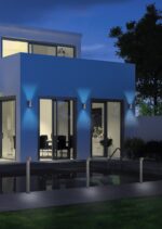 In der Abenddämmerung besticht das moderne zweistöckige Haus von Licht Factory durch große Glasfenster und -türen. Innenleuchten beleuchten den Essbereich, während draußen ein kleiner Pool von sanftem Licht der Roya Outdoor Wandleuchte und einem gepflegten Rasen umgeben ist, was eine einladende Atmosphäre schafft.