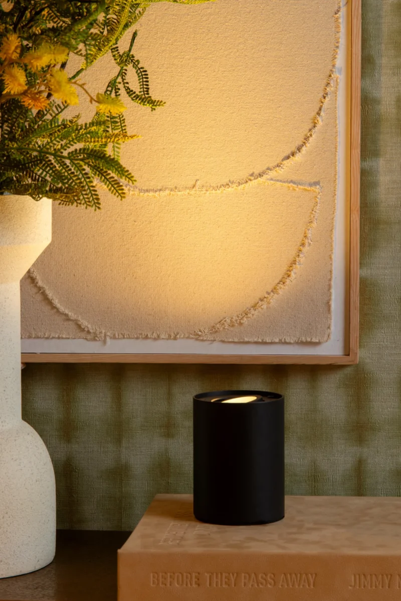 Ein schwarzer Kerzenständer von Licht Factory mit einer brennenden Kerze ziert ein Buch mit dem Titel „BEFORE THEY PASS AWAY“, begleitet von einer Tischleuchte – Tube. Ein strukturiertes Kunstwerk und eine Vase mit Farnen und gelben Blumen werten die grün gemusterte Wand auf.