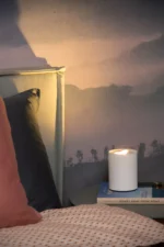 Ein gemütliches Schlafzimmer mit einem weichen rosa Kissen und gemusterten Laken auf dem Bett. Die Tischleuchte - Tube von Licht Factory strahlt auf dem Beistelltisch neben zwei Büchern ein warmes Licht aus. Ein ruhiges, nebliges Landschaftswandbild vervollständigt die Szene im Hintergrund.
