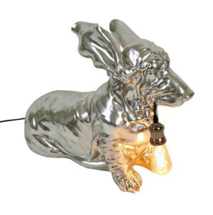 Die „Tischleuchte - Columbo“ ist eine glänzende, metallische Skulptur eines liegenden Hundes, der eine leuchtende Glühbirne im Maul hält. Das Stück ist stark reflektierend und hat eine glatte, glatte Oberfläche, die das Licht wunderbar einfängt.