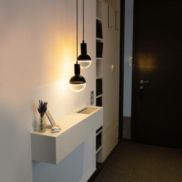 Der minimalistische Flur präsentiert einen schmalen weißen Konsolentisch mit einer kleinen Vase mit Lavendel und einem Bilderrahmen. Darüber erhellen zwei LED-Globe-Pendelleuchten von Licht Factory den Raum. Auf der rechten Seite sind eine dunkle Tür und eingebaute Regale zu sehen.