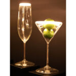 Das LED-Leuchtmittel „Cocktails“ von Licht Factory beleuchtet ein hohes Champagnerglas und ein Martiniglas mit Limetten wunderschön und erzeugt einen warmen, leuchtenden Effekt vor einem dunklen Hintergrund.