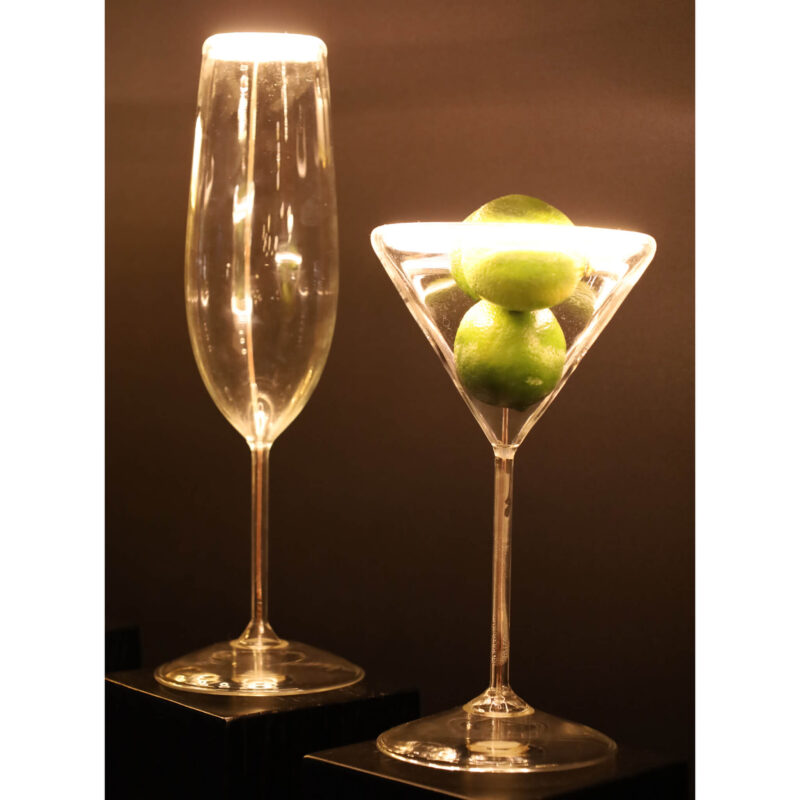 Das LED-Leuchtmittel „Cocktails“ von Licht Factory beleuchtet ein hohes Champagnerglas und ein Martiniglas mit Limetten wunderschön und erzeugt einen warmen, leuchtenden Effekt vor einem dunklen Hintergrund.