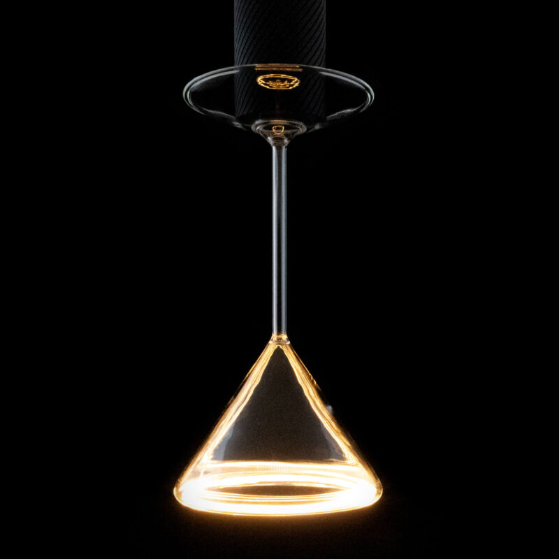 Das LED-Leuchtmittel - Cocktails von Licht Factory hängt kopfüber und strahlt aus seinem Sockel ein warmes gelbes Licht aus. Das leuchtende Weinglas erzeugt vor dem dunklen Hintergrund einen sanften Halo-Effekt.
