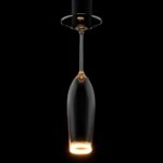 Ein LED-Leuchtmittel - Cocktailglas von Licht Factory schwebt in der Dunkelheit, wobei sein Boden warm leuchtet und einen Halo-Effekt erzeugt. Der Stiel reflektiert das Licht subtil.