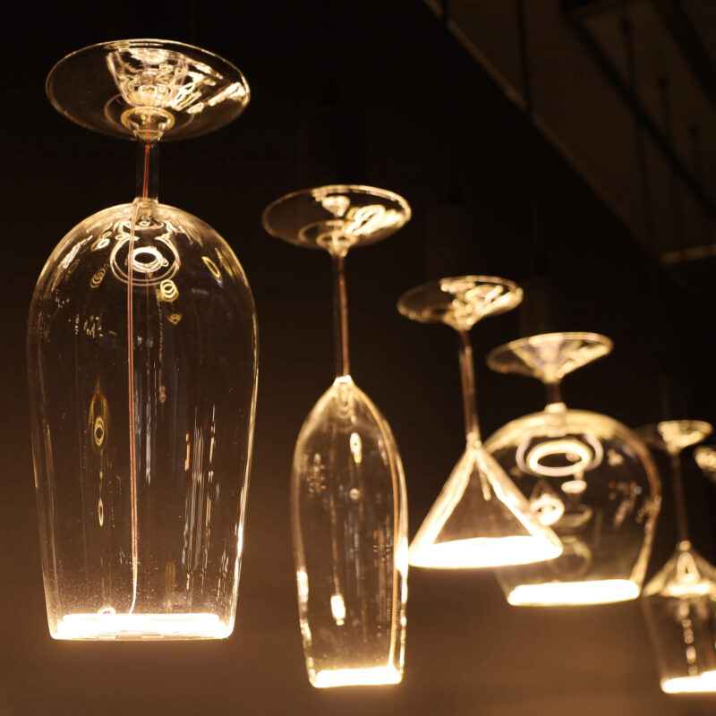 Die LED-Leuchtmittel - Cocktails von Licht Factory beleuchten verschiedene Arten von Glaswaren, wie Wein- und Martinigläser, die kopfüber aufgehängt sind. Die Lichter werfen einen warmen Schein auf einen dunklen Hintergrund.