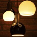 Nahaufnahme von drei hängenden LED-Pendelleuchten „Billard“ von Licht Factory vor einer Ziegelwand, mit kugelförmigen Glasschirmen in warmem Gelb und glänzendem Schwarz. Der schwarze Schirm ist unten offen und sorgt für eine sanfte Beleuchtung der darunterliegenden Fläche.