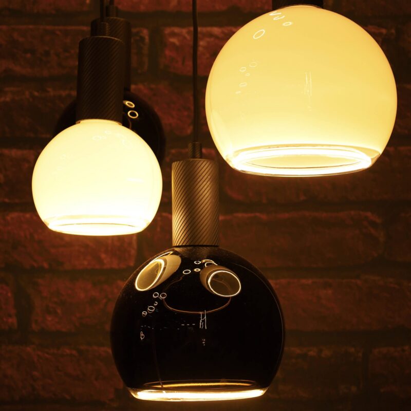 Nahaufnahme von drei hängenden LED-Pendelleuchten „Billard“ von Licht Factory vor einer Ziegelwand, mit kugelförmigen Glasschirmen in warmem Gelb und glänzendem Schwarz. Der schwarze Schirm ist unten offen und sorgt für eine sanfte Beleuchtung der darunterliegenden Fläche.